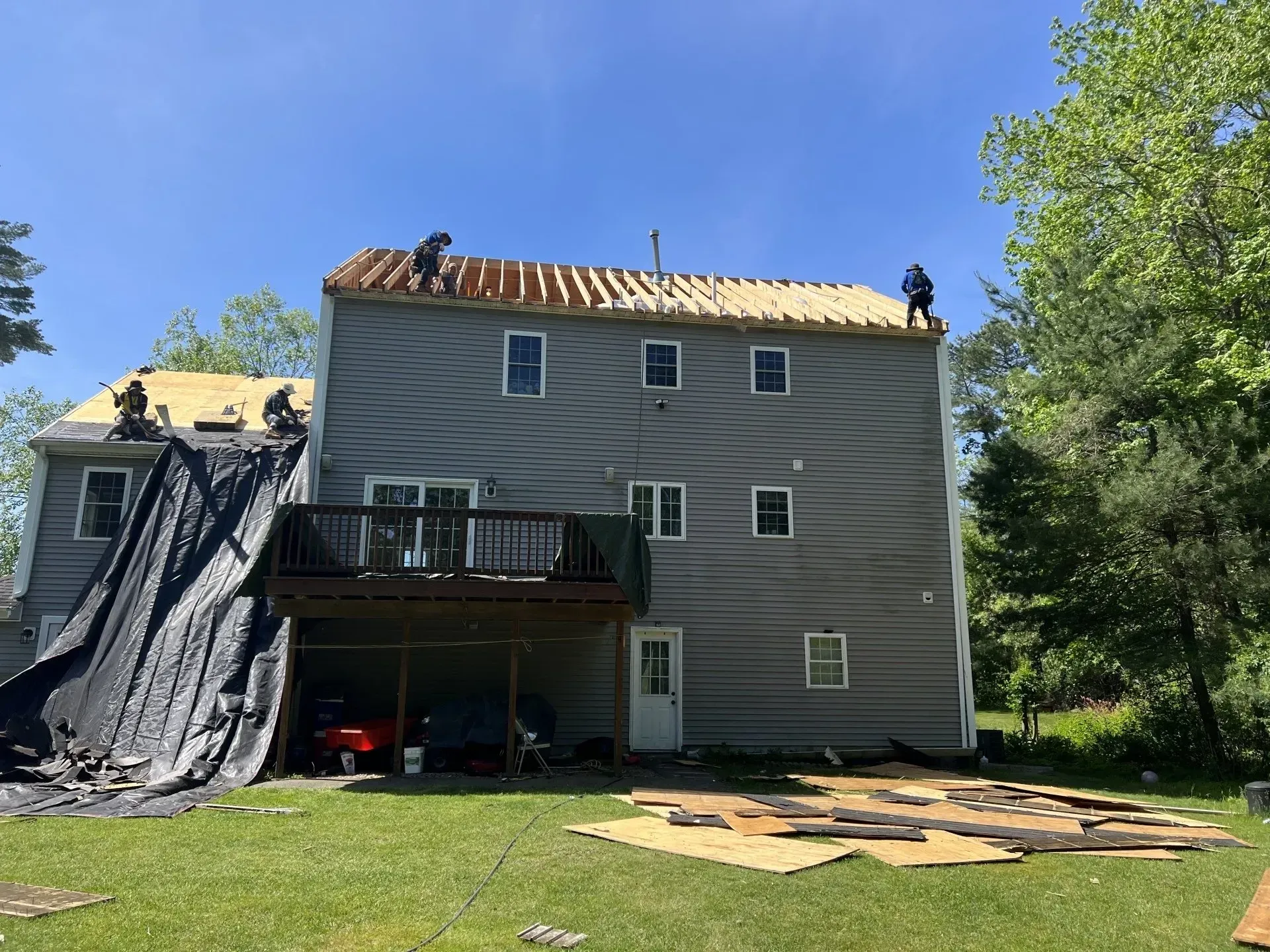 Roofers working on a two-story house, with some roof sections exposed. Blue sky and trees surround the building.