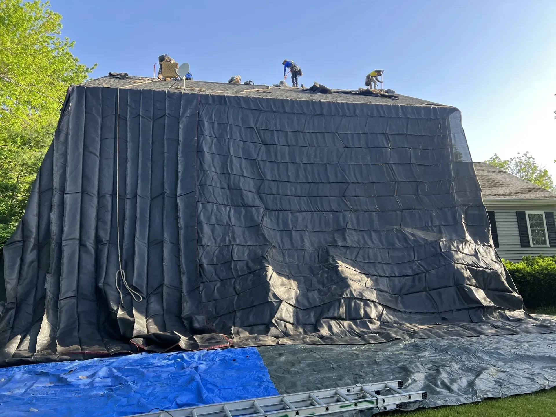 Roofers working on a house covered with a large black tarp. Blue tarp and ladder are on the ground.