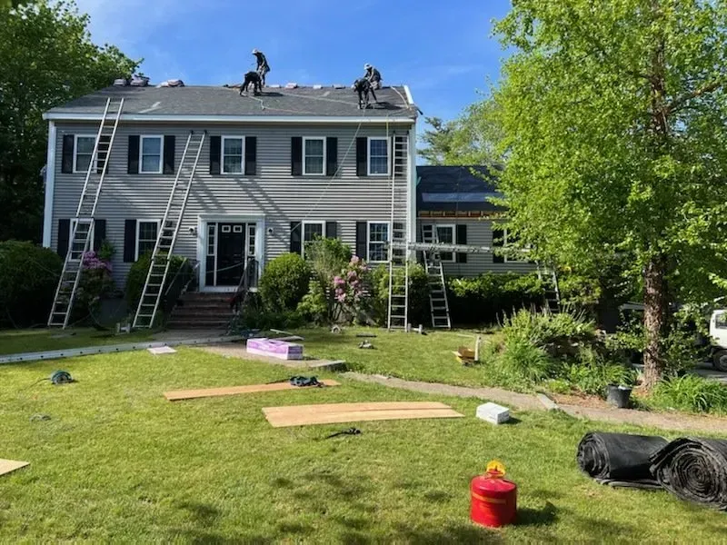 Roofers working on a two-story gray house. Ladders propped against the roof and walls; green grass in the yard.