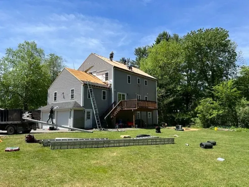 Workers on a roof, replacing shingles on a two-story gray house. Ladders, tools, and materials on lawn.