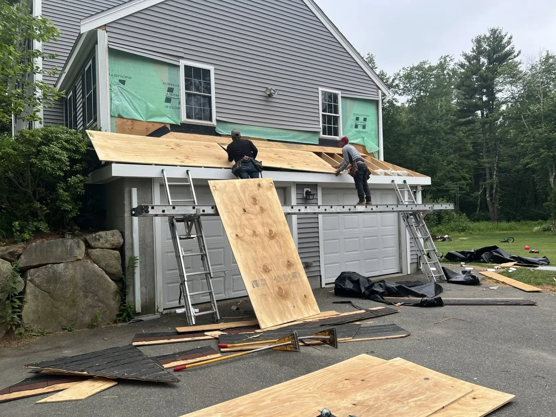 Roofers working on a house with gray siding. Men installing plywood, asphalt shingles, and standing on ladders.