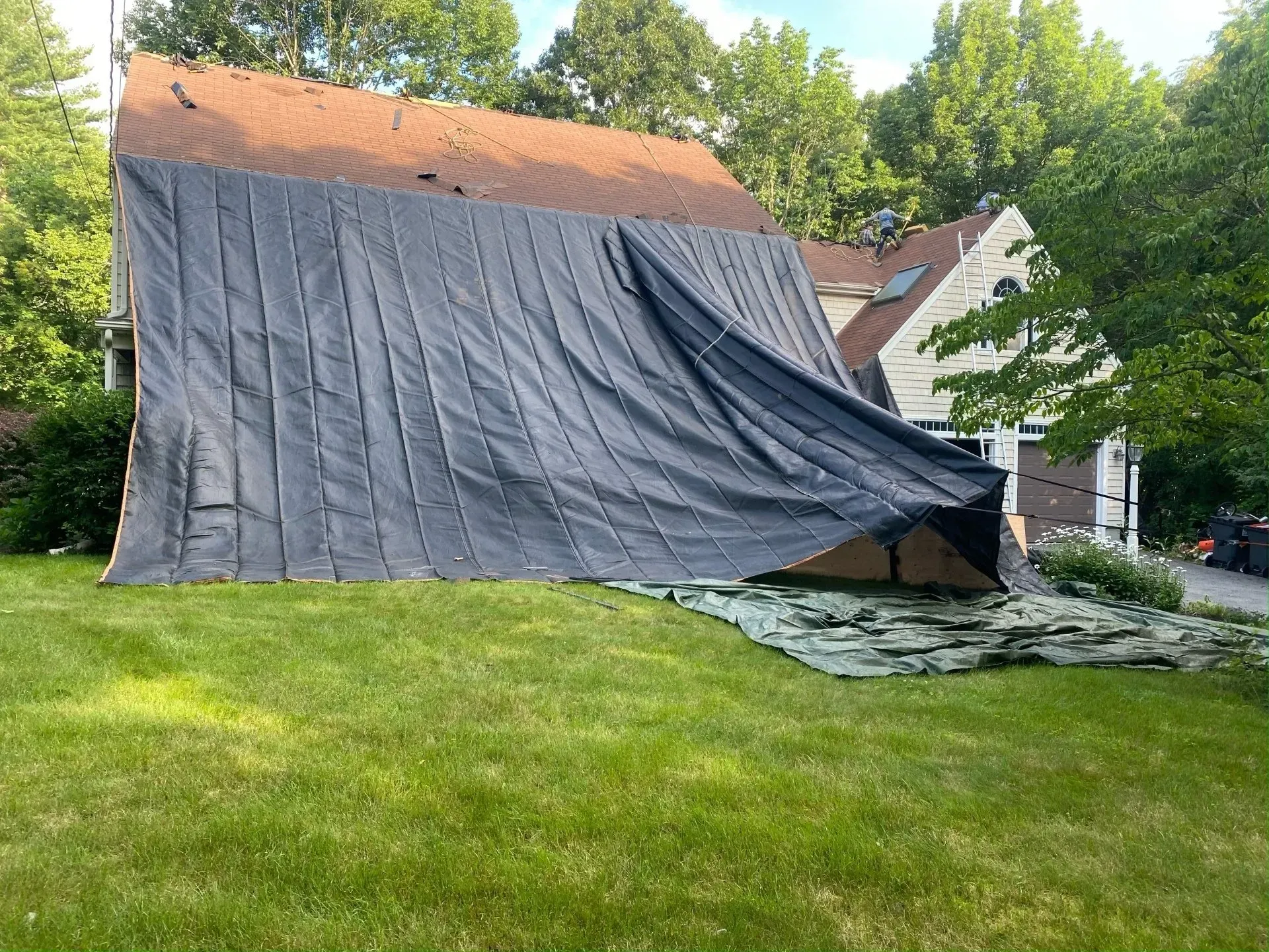 Black tarp covers damaged roof of a house, billowing in the wind. Green grass in the yard.