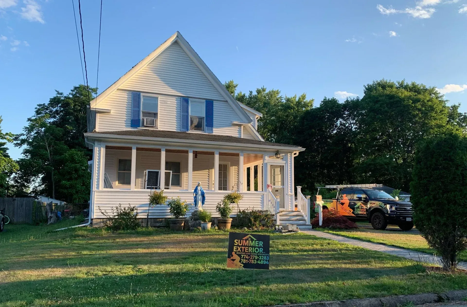 White house with blue shutters, covered porch, and a sign on the lawn. A truck is parked to the right.
