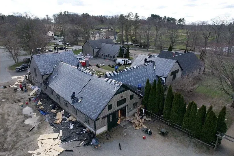 Aerial view of a building with a damaged roof, likely under construction. Gray roof and wooden materials visible.