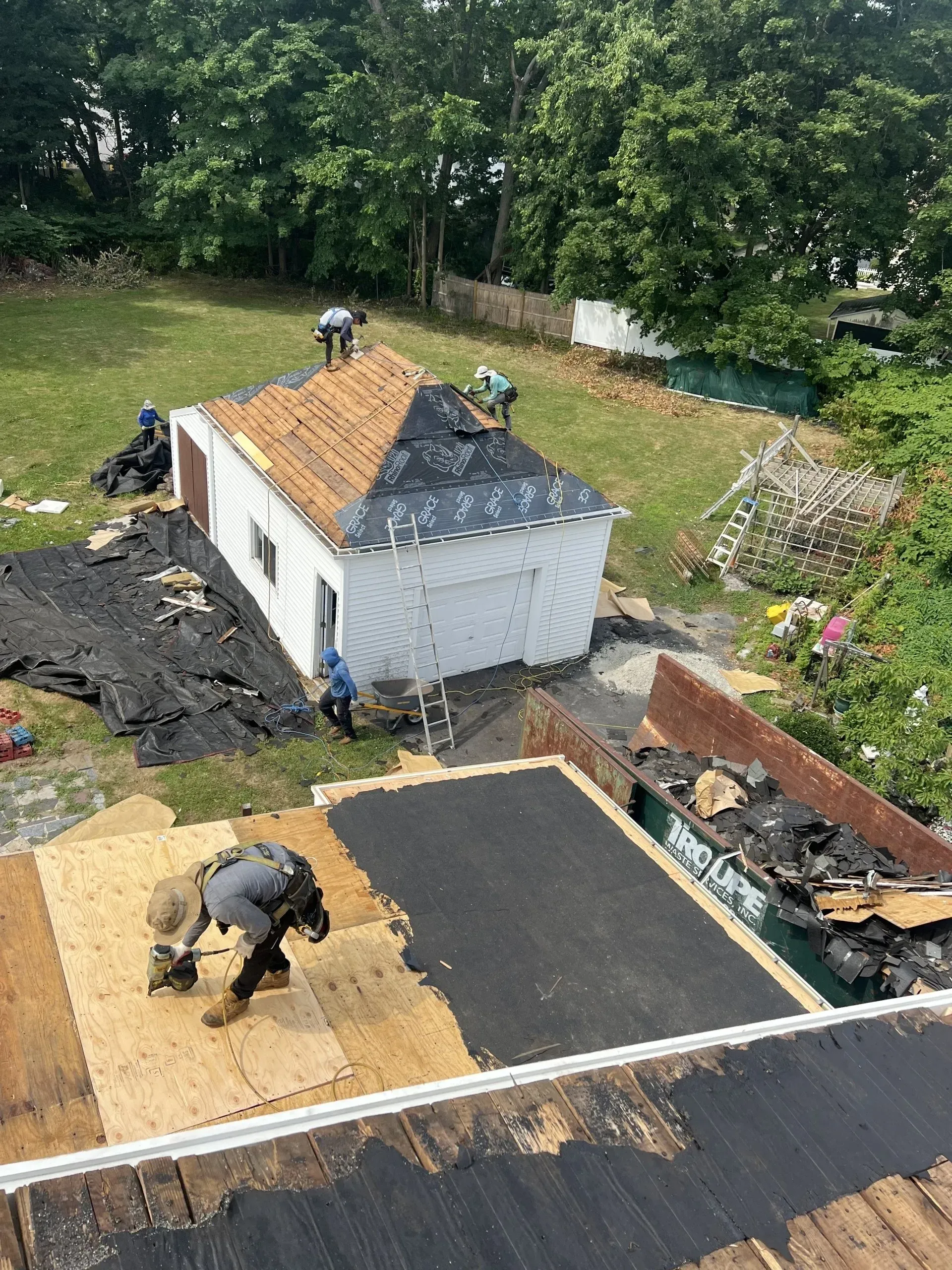 Roofers working on a building, applying shingles. Black and brown roof, green grass, trees, and blue sky.