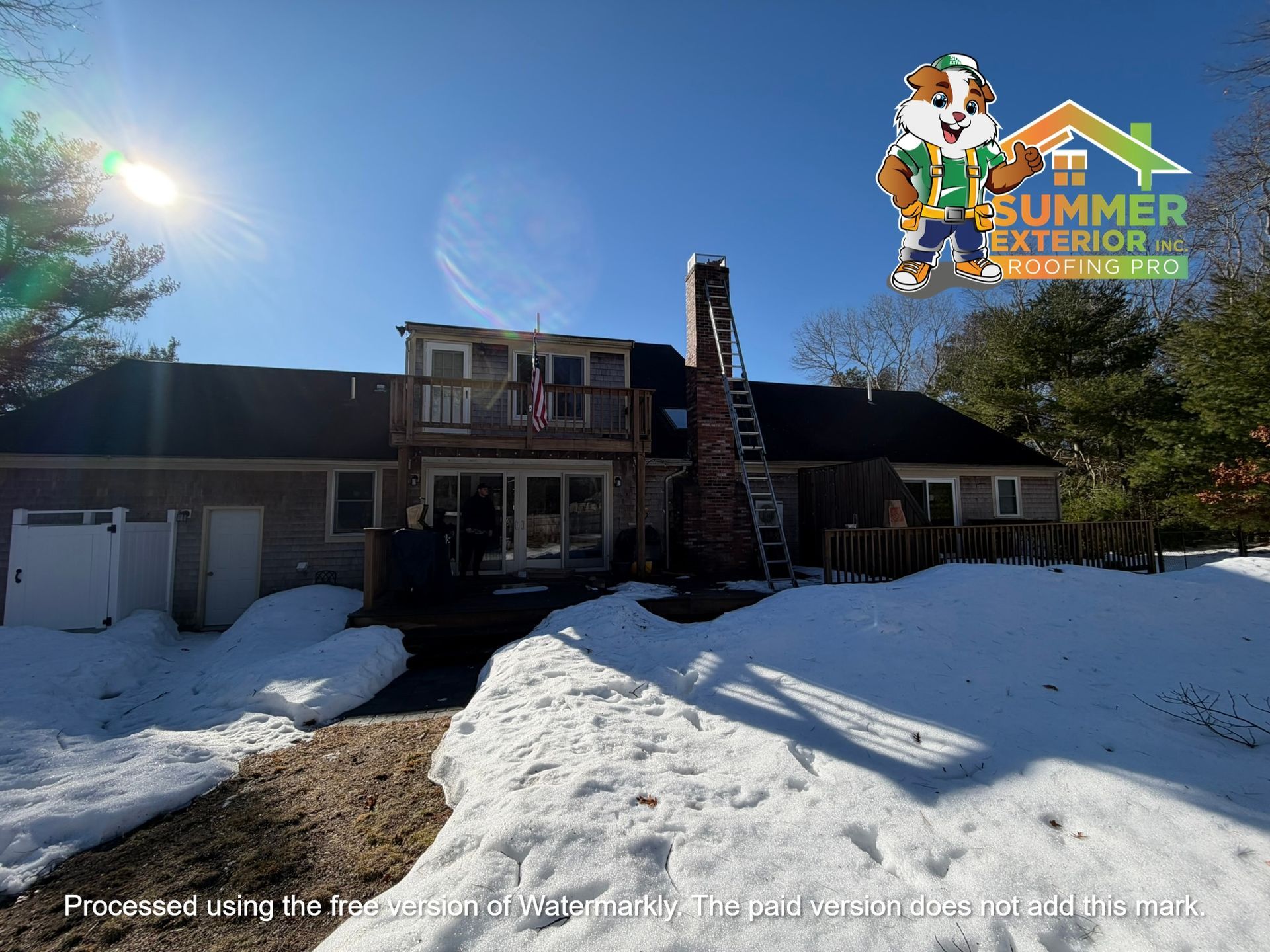 A snowy backyard view of a house with a brick chimney, a balcony, and a ladder leaning against the roof.