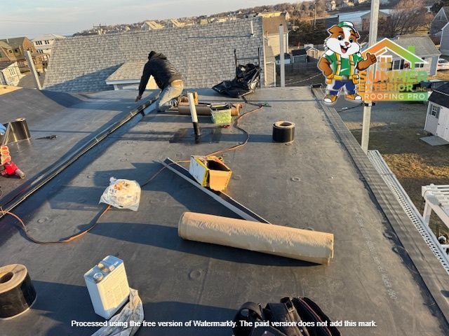 Roofer working on a flat, black roof. Equipment, including a roll of material, is scattered about. Daylight setting.