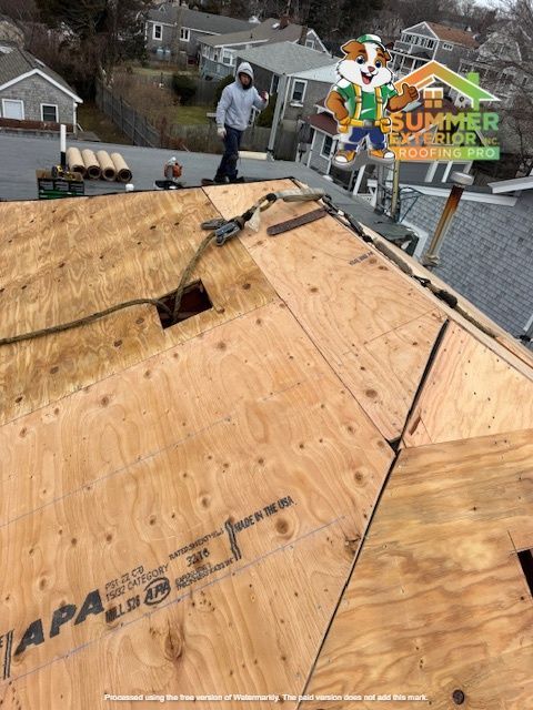 Roofer on a plywood roof, with rolls of material nearby. Building in background.