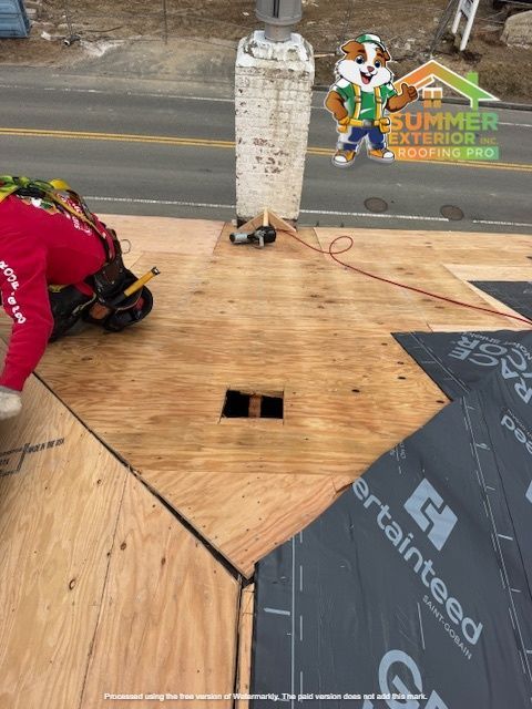 Roofer on a roof installing shingles. Chimney, plywood, and a cartoon dog logo are visible.