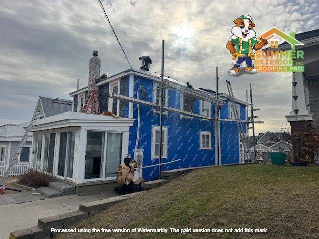 Construction workers installing siding on a two-story house, blue wrap visible. Scaffolding, sunny day, residential setting.