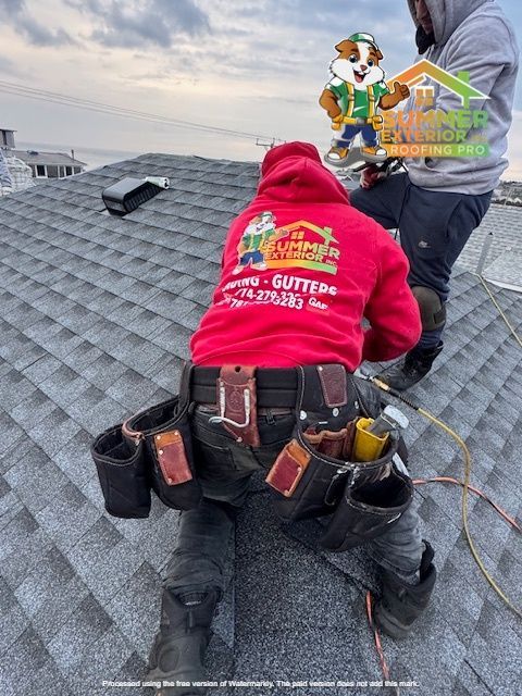 Two roofers working on a gray shingled roof, one in a red hoodie and tool belt.