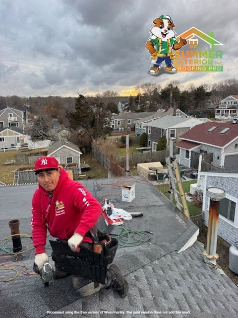 Roofer in red hoodie on a rooftop, holding a nail gun, with cloudy sky and houses in the background.