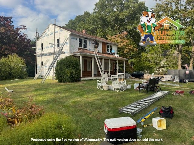 House exterior renovation in progress; workers on ladders, green yard, tools visible.