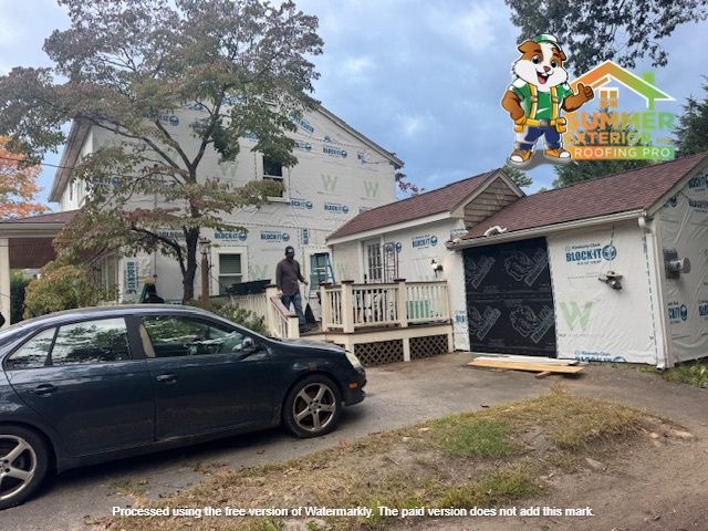 A house and garage are under construction, covered in white wrap. A car is parked in the driveway. A person is on the porch.