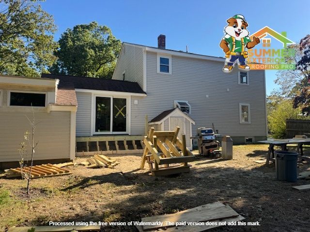 Exterior house under construction with light gray siding, wooden deck, and a roof, sunny day.