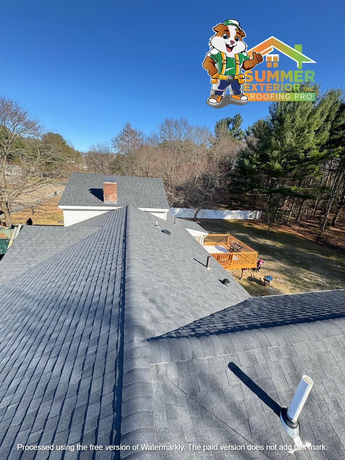 An aerial view of a dark grey shingled residential roof, featuring a company logo and a plumbing vent pipe.