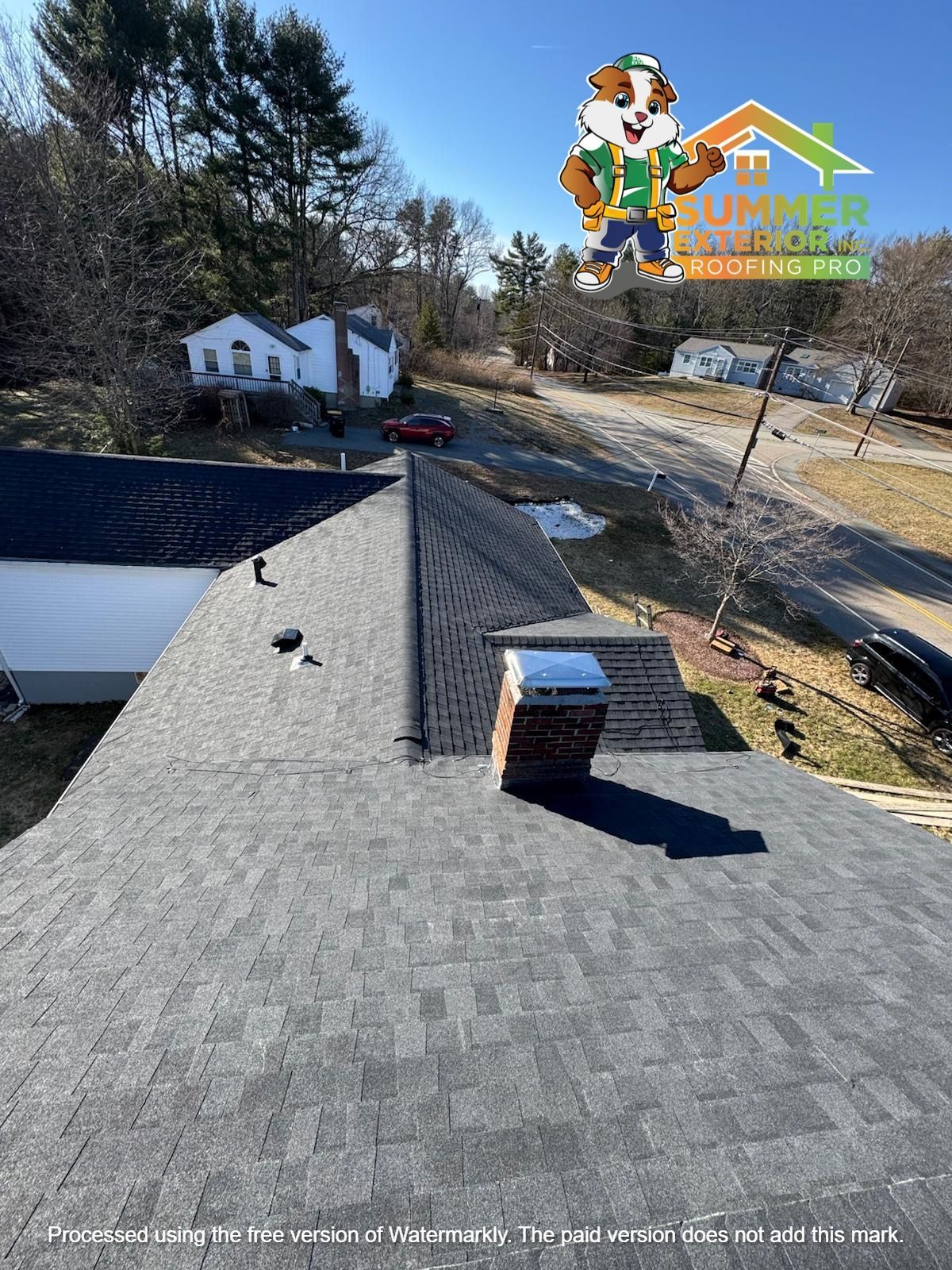 A newly shingled gray roof with a brick chimney under a clear blue sky, featuring a company logo in the upper right.