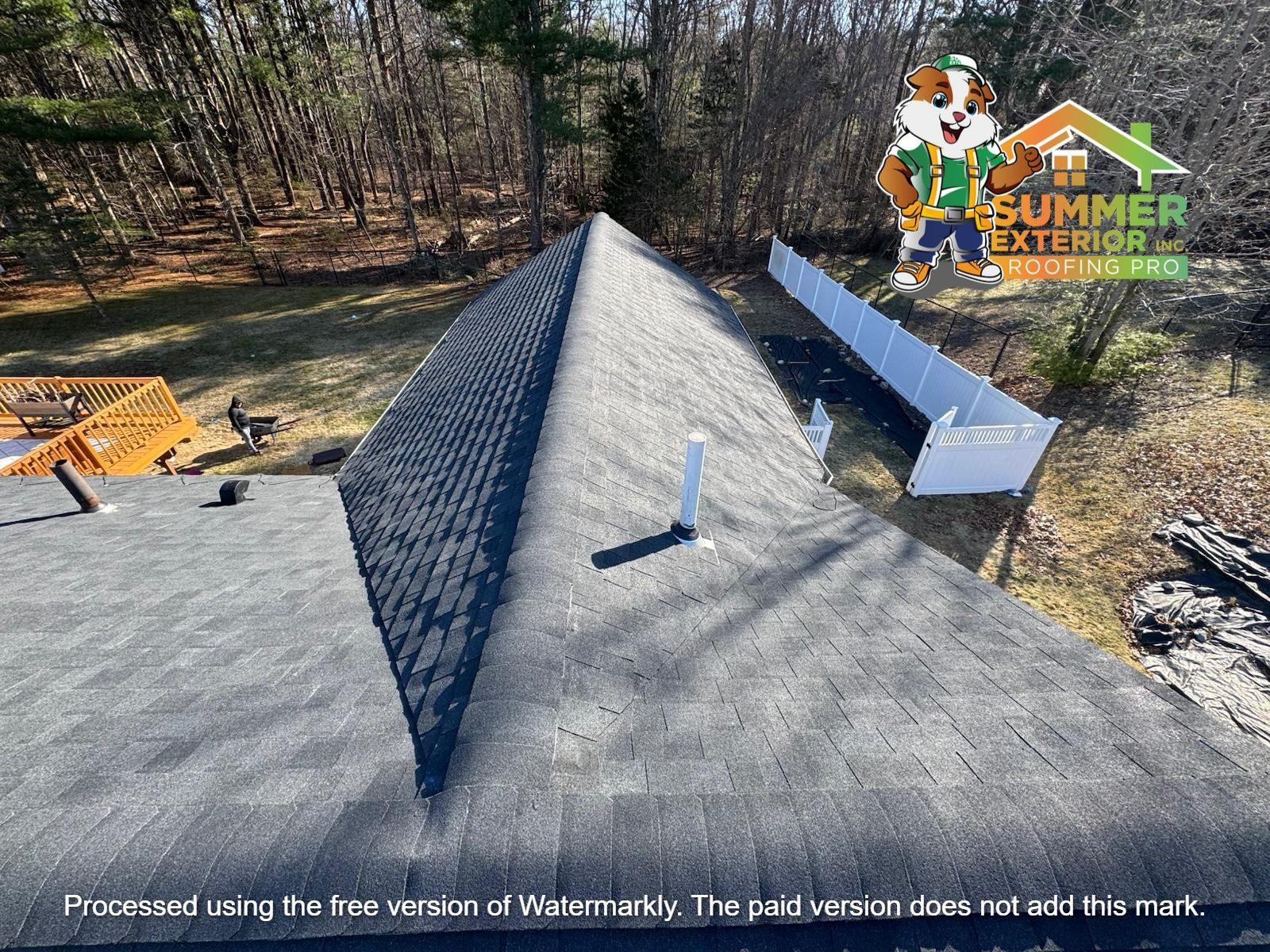 A drone-view of a grey asphalt shingle roof with a central ridge, a white vent pipe, and a white fence in the background.