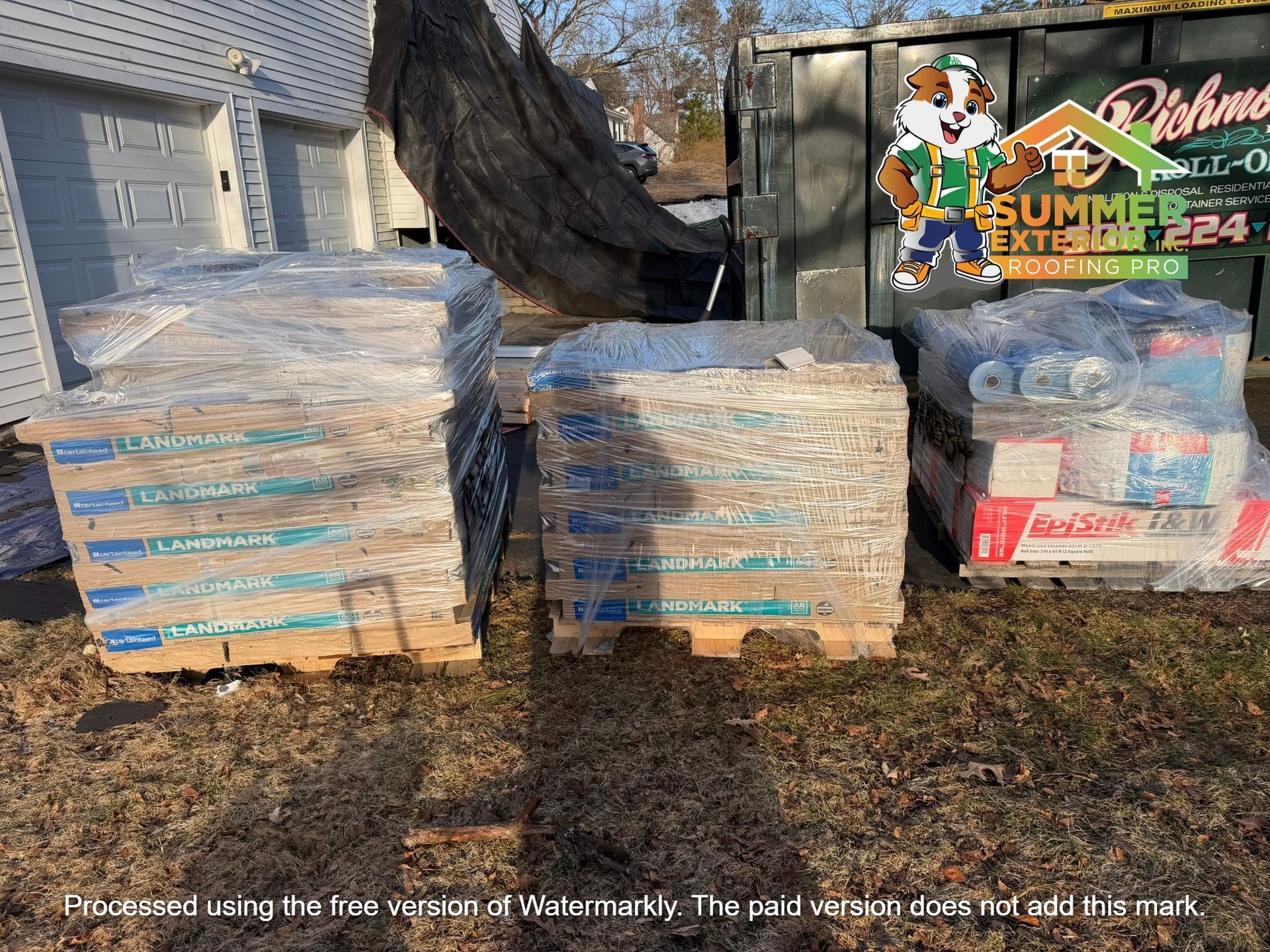 Three pallets of building materials, including wrapped insulation and shingles, sit on a lawn near a dumpster.