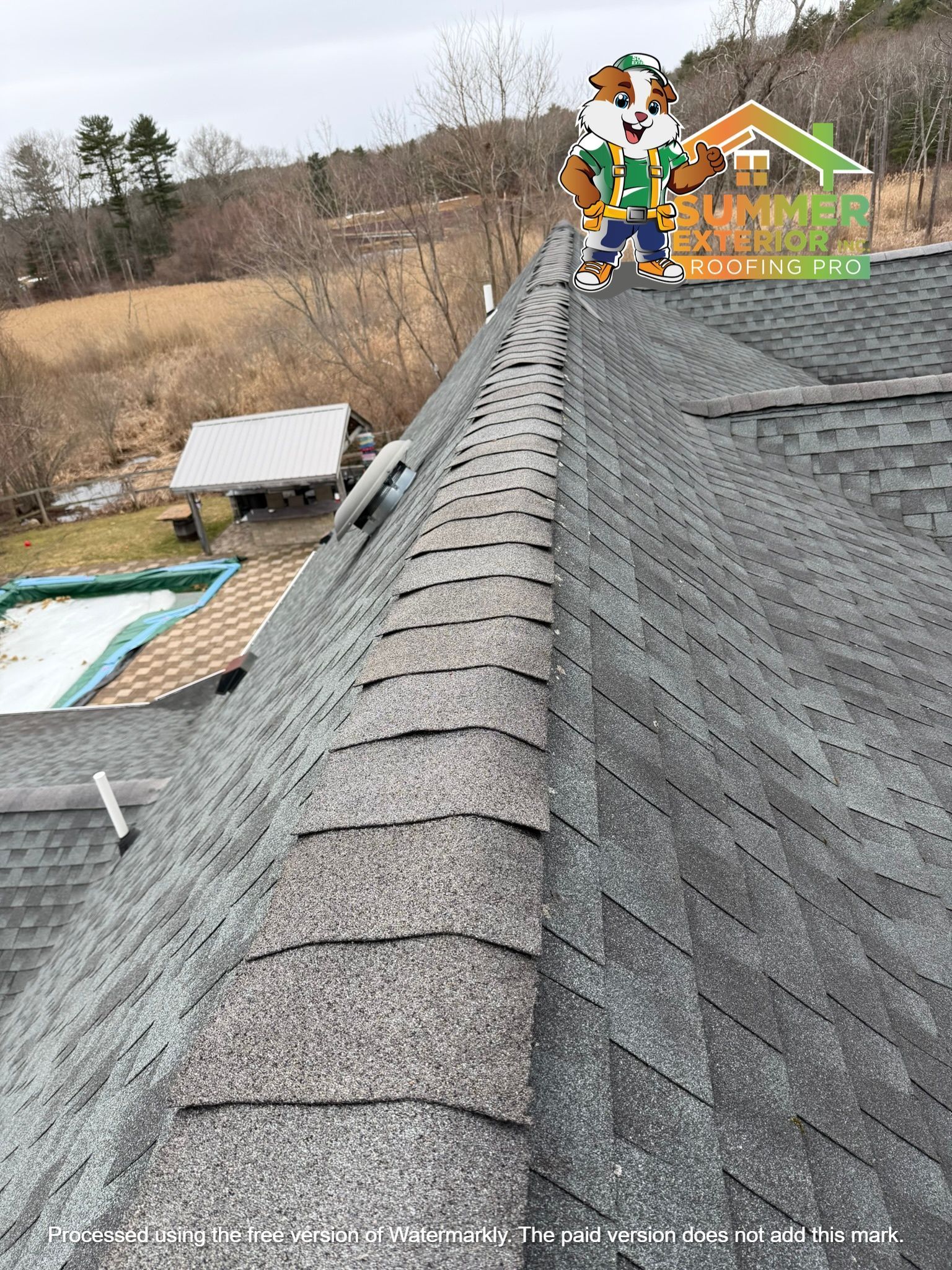A high-angle view of a gray asphalt shingle roof ridge with a mascot character logo overlay in the top right corner.