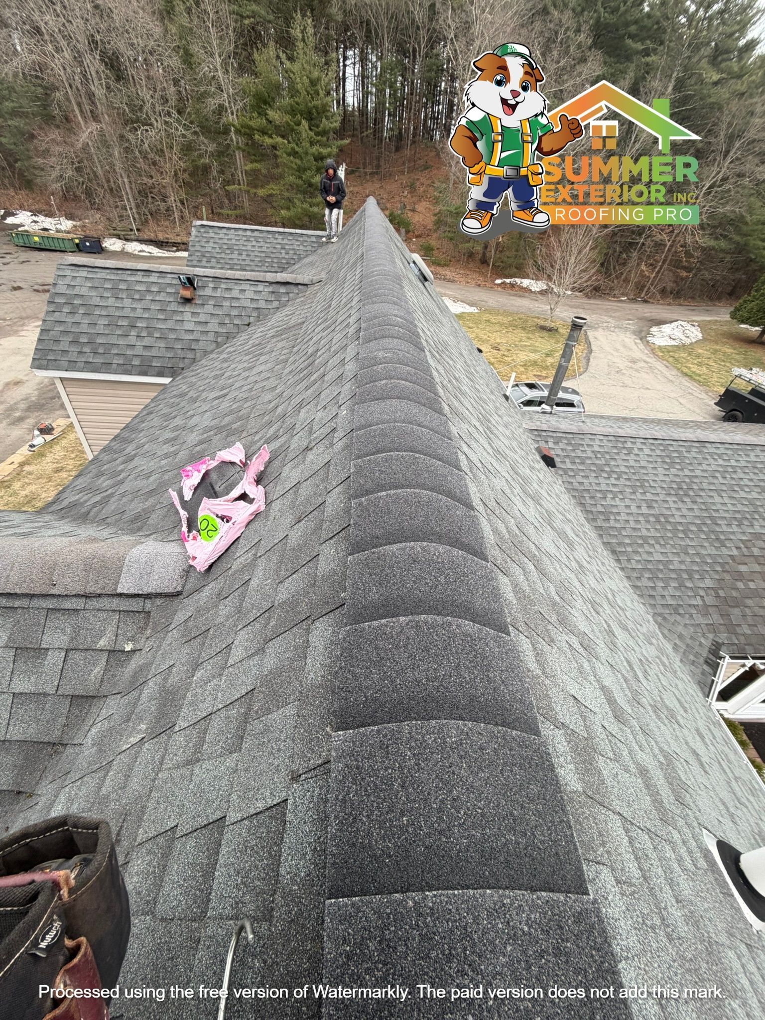 A view from a rooftop looking along the ridge of newly installed asphalt shingles, with a worker visible in the distance.