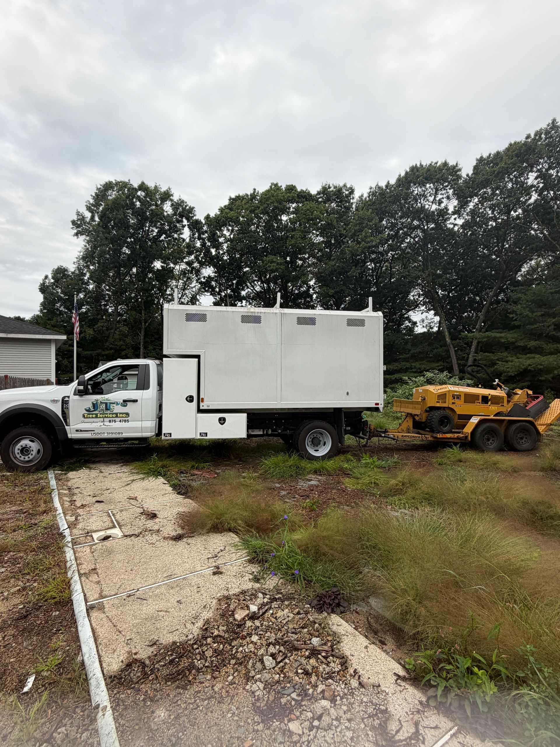 White utility truck and yellow skid steer on a muddy driveway beside a house and trees