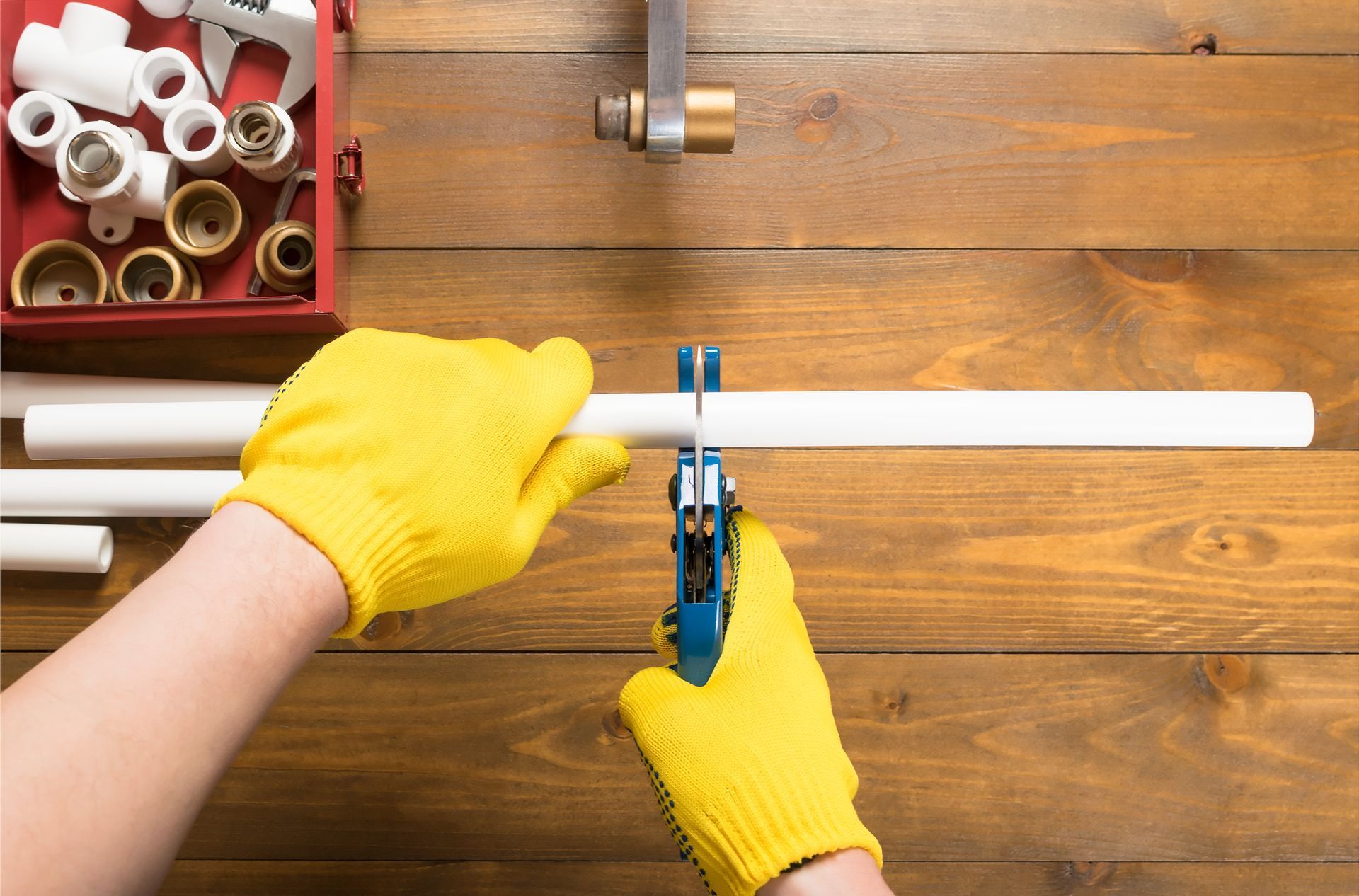Person with yellow gloves cutting white PVC pipe with a blue cutter on a wooden surface.