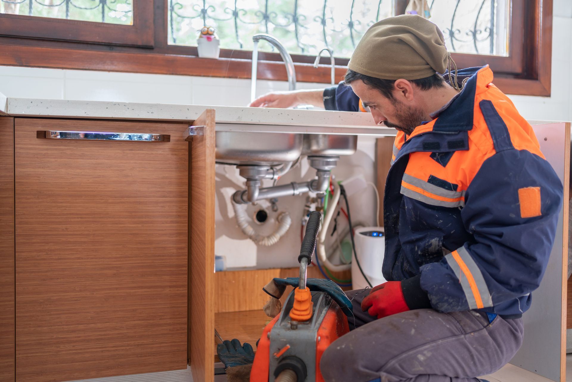 Plumber kneeling by a kitchen sink, working on the pipes with tools; wearing a blue and orange uniform.