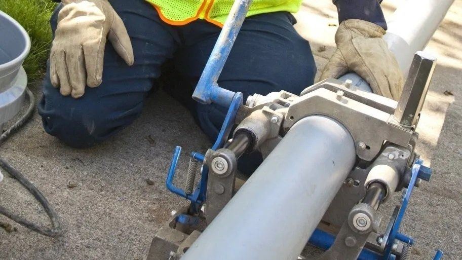 Person in gloves using a pipe fusion machine to join two gray pipes outdoors on a concrete surface.