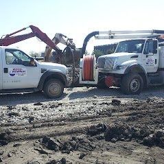 Two white trucks with equipment, one with an excavator arm, working in a dirt lot.