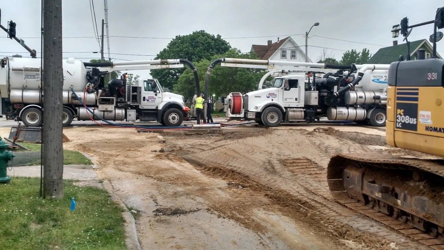 Two white trucks and an excavator on a muddy road; construction work happening.