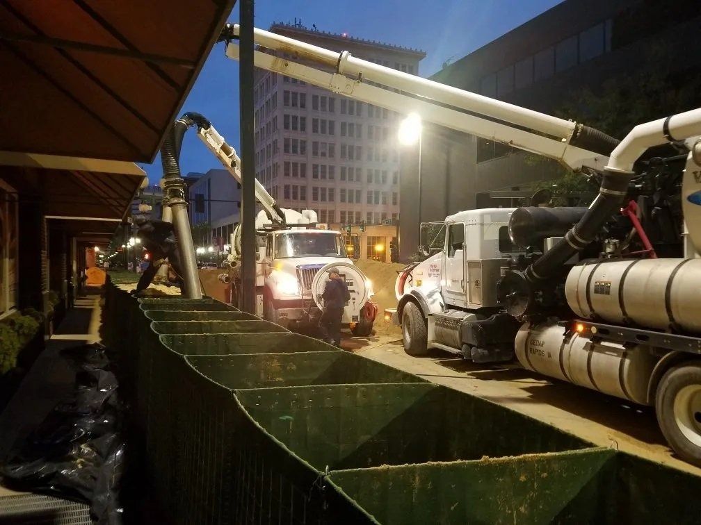 Construction site at night. Two trucks with long arms, digging near a building, with a green barrier in front.