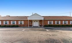 Red brick building with white trim, windows, and a central entrance under a white awning; paved lot in front.