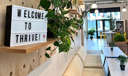Welcome to Thrive! sign above a bench in a bright, modern office with plants.