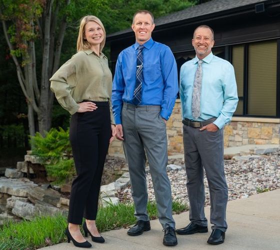 Three people stand outside a building, smiling in business-casual attire.