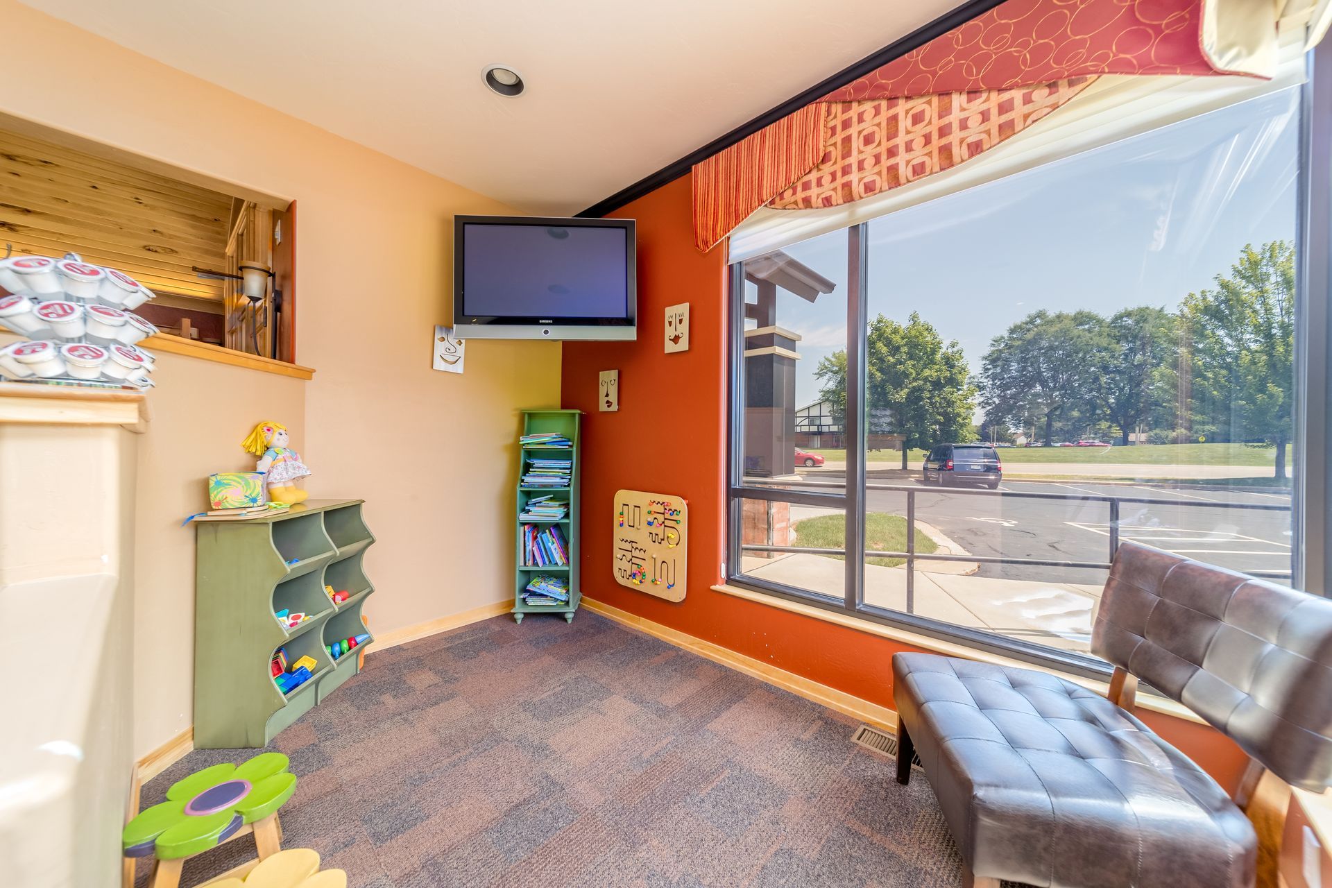 Bright kids’ playroom with TV, toy shelves, and a large window overlooking a parking lot.