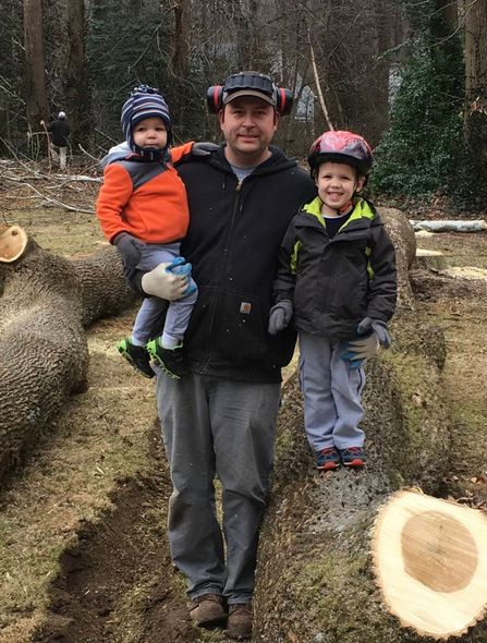 A man holds two children in his arms beside a tree stump.