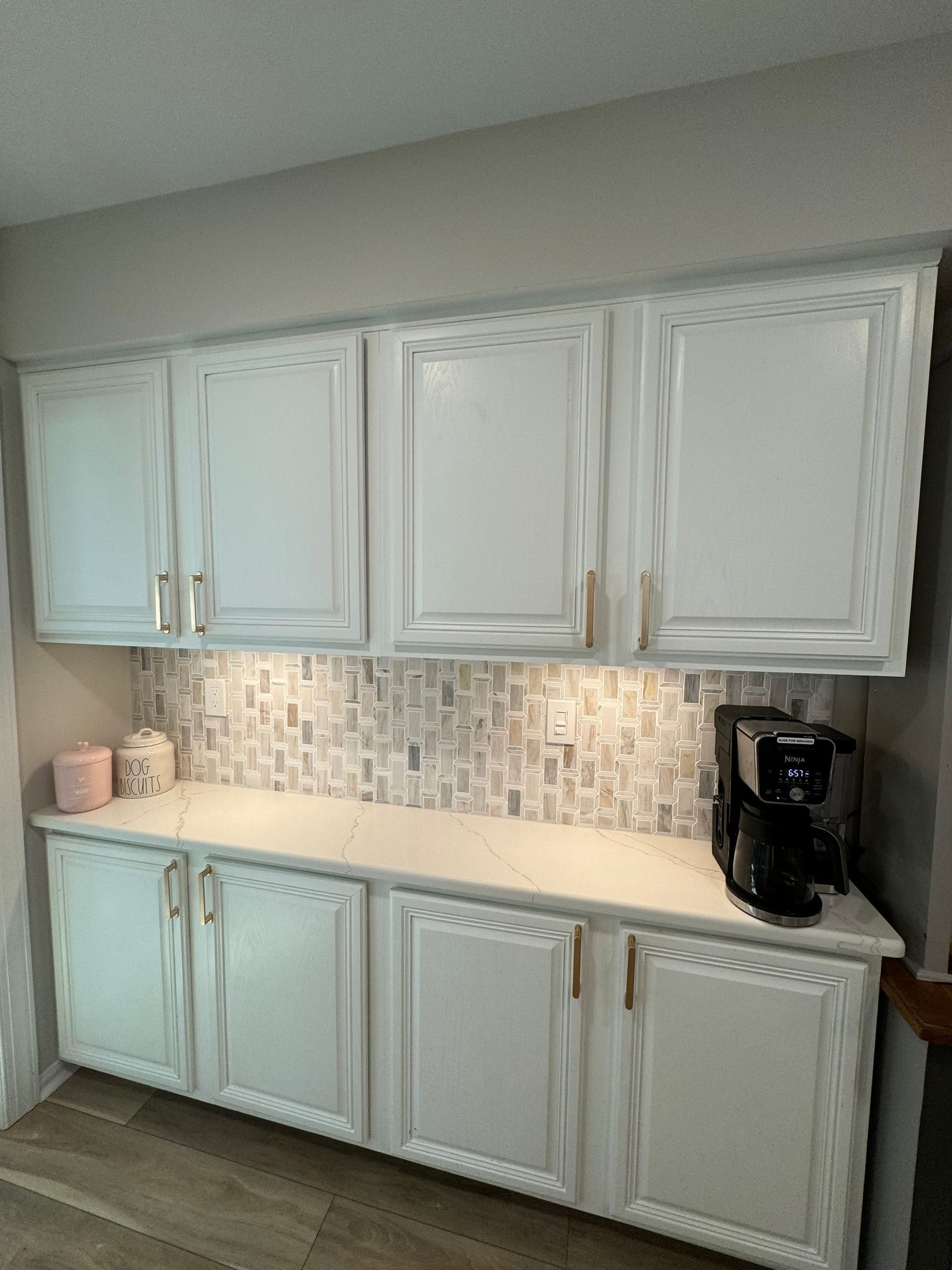 White cabinets above and below a counter with coffee maker. Tile backsplash, gold hardware, and lighting.
