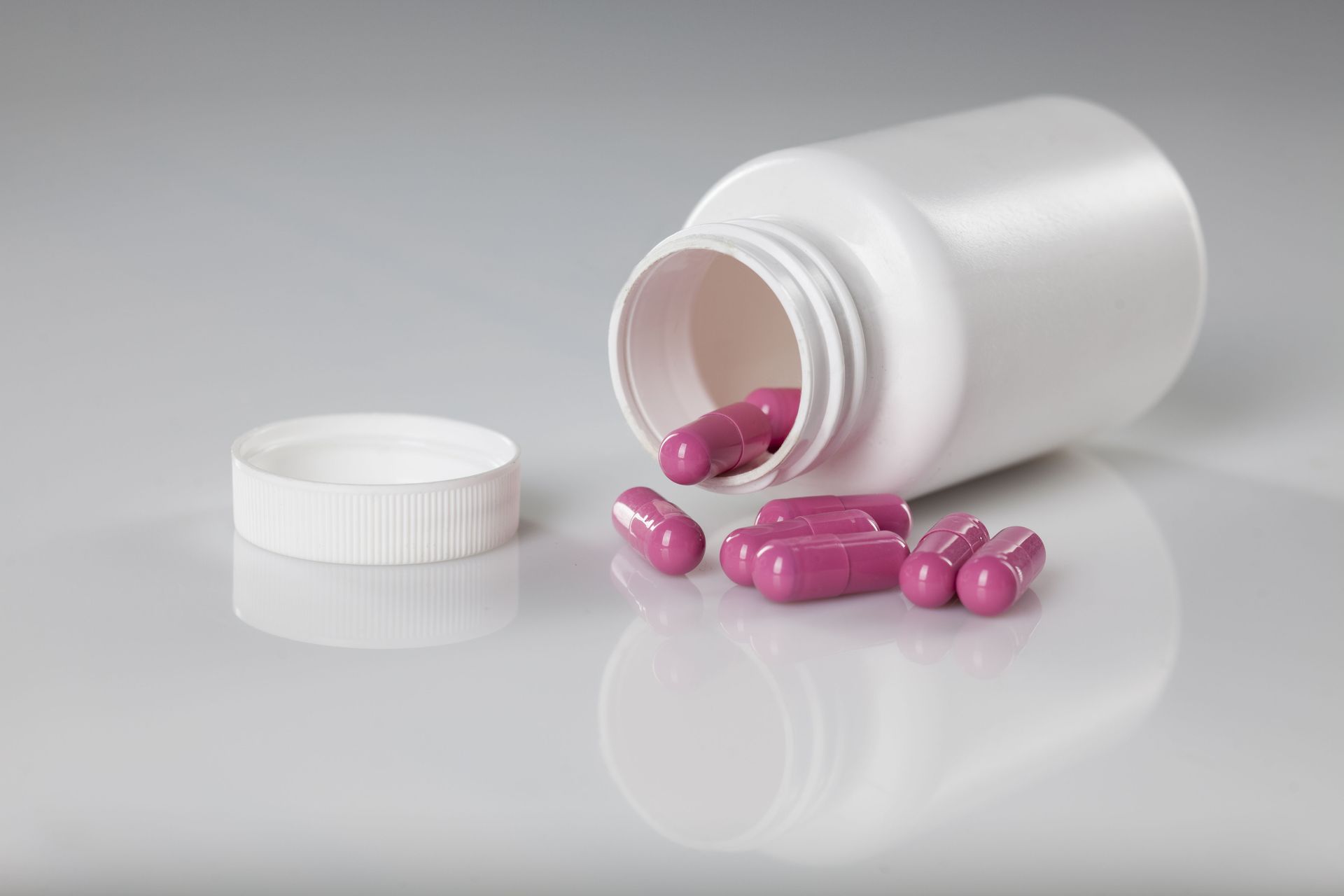 White pill bottle tipped over, spilling pink capsules onto a reflective surface.