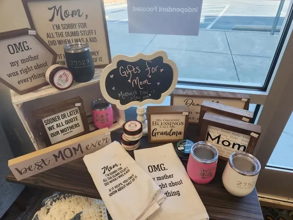 Mother's Day gifts on display at a store, including signs, mugs, and towels, near a window.