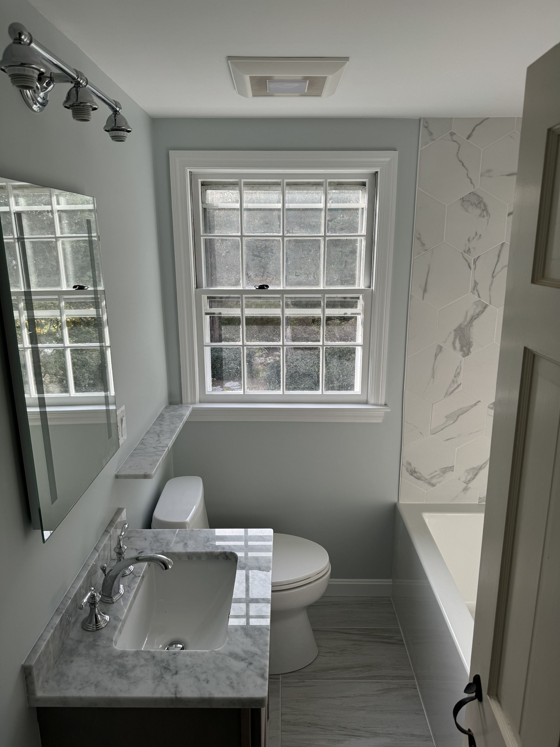 A bathroom with a toilet , sink , mirror and window.