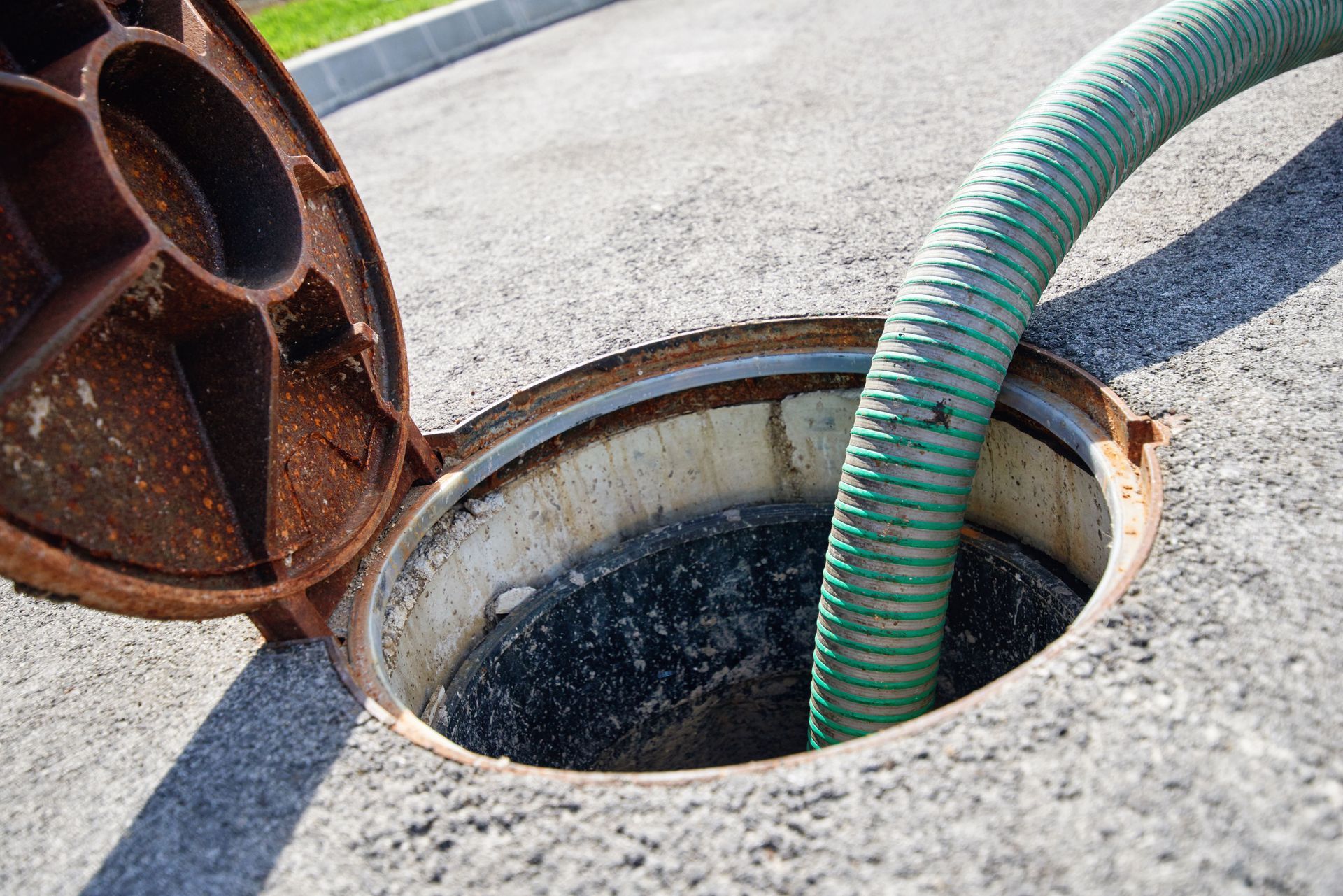 Open manhole with a green hose inside, on asphalt. Rusty lid is open.