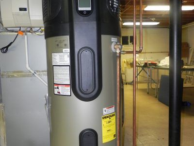 Large gray water heater in a utility room with pipes, labels, and an open doorway to a workspace.