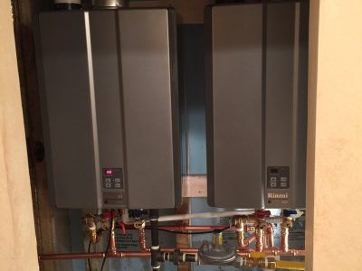 Two gray wall-mounted boilers with exposed copper pipes in a utility closet
