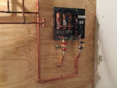 Copper piping and an open wall-mounted HVAC unit installed on plywood backing.