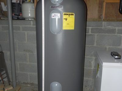 Gray water heater tank in a basement with cinderblock walls and a yellow warning label.