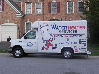 White service van with “Water Heater Services” parked on a street in front of brick houses.