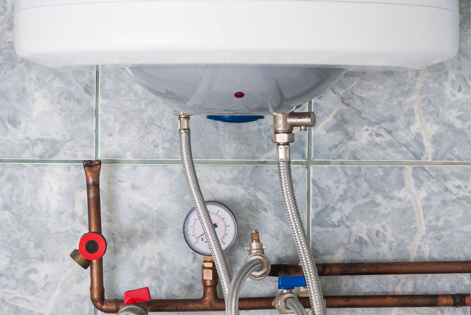 Water heater with exposed pipes, valves, and hoses against a tiled bathroom wall.