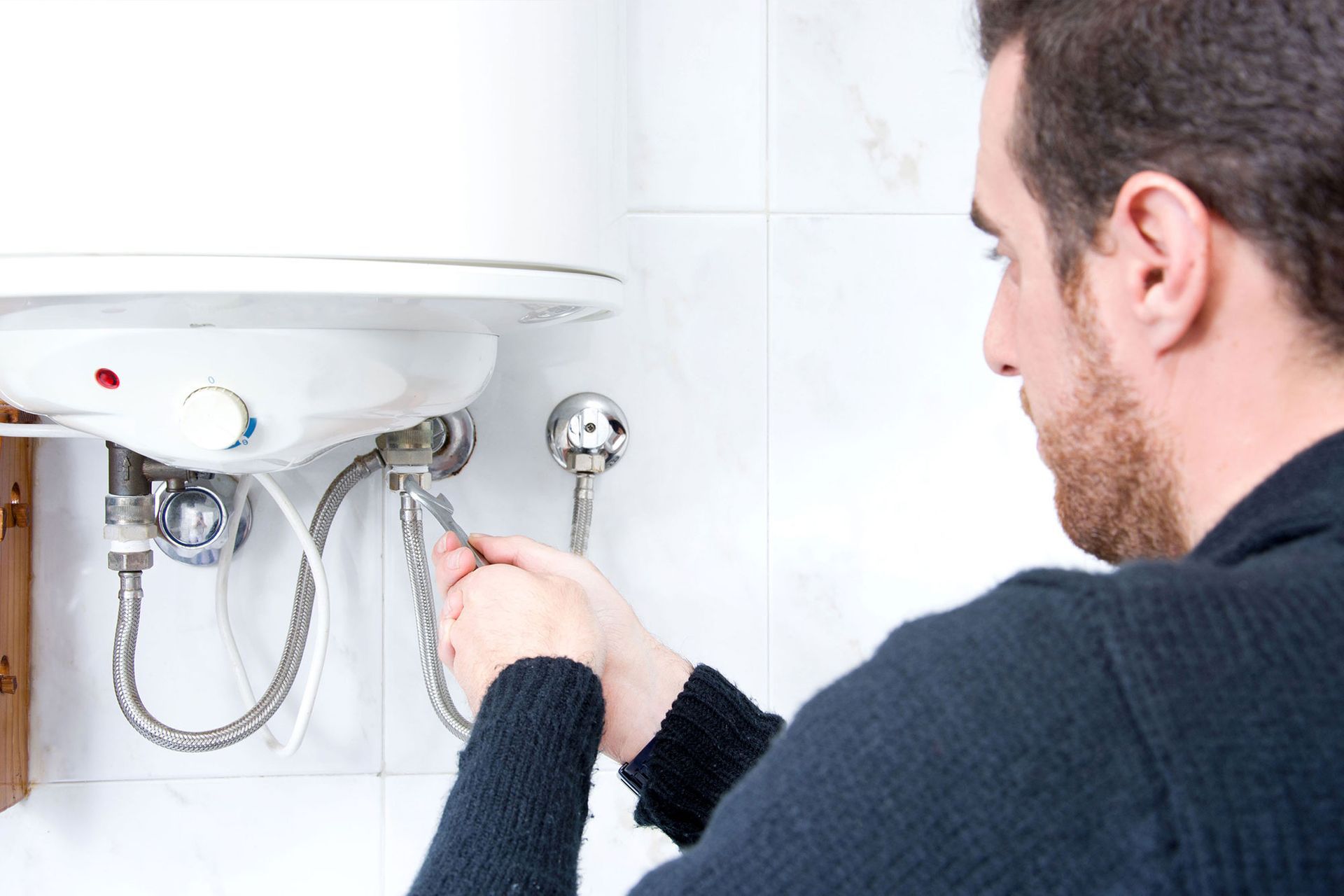 Man repairing a wall-mounted water heater with a wrench in a tiled bathroom