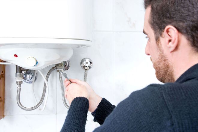 Man repairing a wall-mounted water heater with a wrench in a tiled bathroom